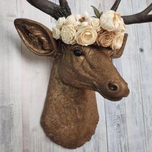 Faux taxidermy deer head that's been painted antique gold with chestnut brown accenting. The antlers are chestnut brown and atop the deers head is a cream floral crown made out of wooden sola flowers