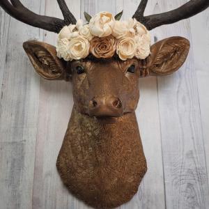 Faux taxidermy deer head that's been painted antique gold with chestnut brown accenting. The antlers are chestnut brown and atop the deers head is a cream floral crown made out of wooden sola flowers