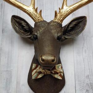 Faux taxidermy deer head, painted chestnut brown with gold accents. The antlers are gold and there is a multi colored brown tonal bow tie on the deers neck