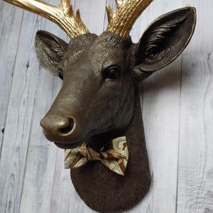 Faux taxidermy deer head, painted chestnut brown with gold accents. The antlers are gold and there is a multi colored brown tonal bow tie on the deers neck