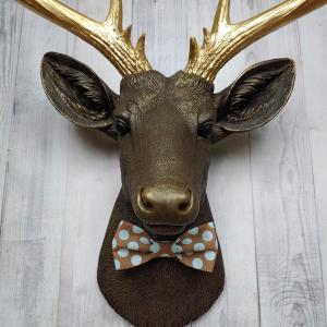 Faux taxidermy deer head. Painted chestnut brown with gold accents and gold antlers. There is a blue and brown polka dot bow tie on the deers neck.