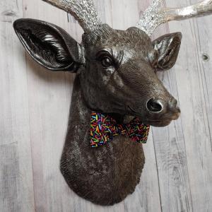 Faux taxidermy deer head painted rubbed bronze with silver accents and silver antlers. The deer has a bow tie on it's neck in black with colorful sprinkles.