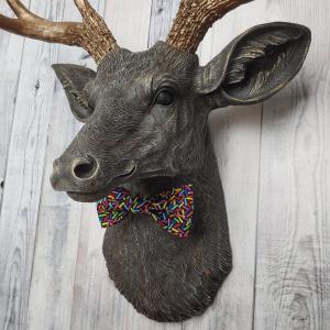 Faux taxidermy dee head painted chestnut brown and accented with antique white and antique gold. The antlers have been painted antique gold and the eyes have been painted black. A fabric bow tie that has a black background with multi colored sprinkles accents the deers neck.