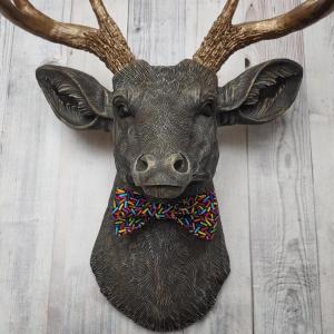 Faux taxidermy dee head painted chestnut brown and accented with antique white and antique gold. The antlers have been painted antique gold and the eyes have been painted black. A fabric bow tie that has a black background with multi colored sprinkles accents the deers neck.