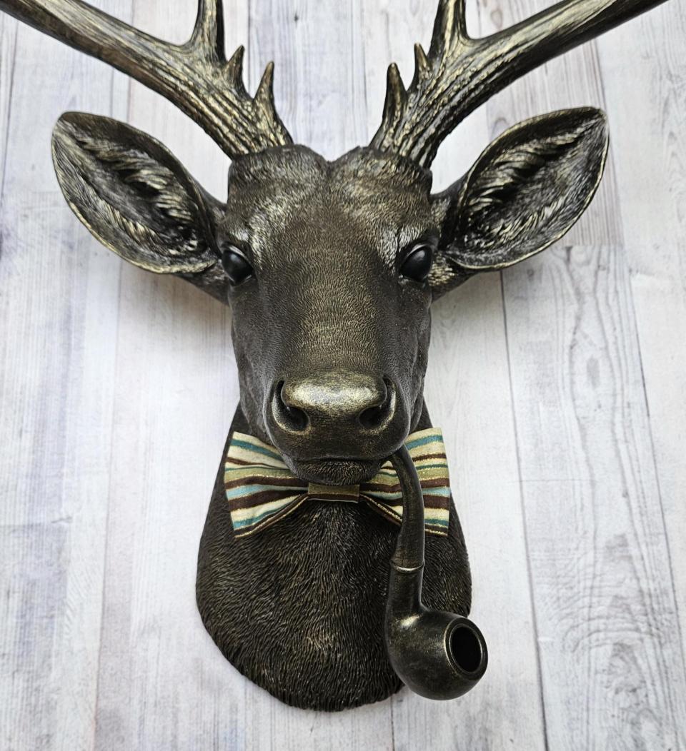 faux taxidermy deer head, rubbed bronze with gold accents. A smoking pipe hangs from the deers mouth and its wearing a striped bow tie