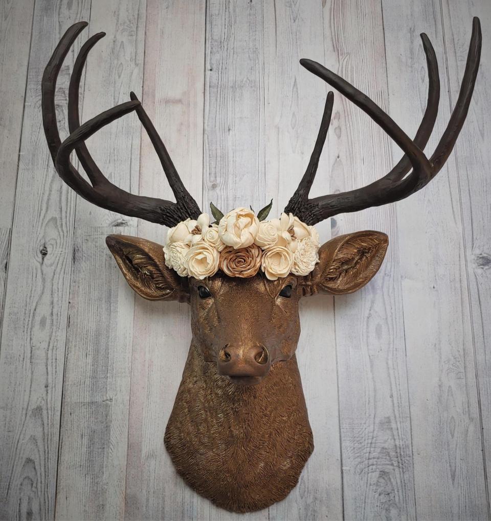 Faux taxidermy deer head that's been painted antique gold with chestnut brown accenting. The antlers are chestnut brown and atop the deers head is a cream floral crown made out of wooden sola flowers.
