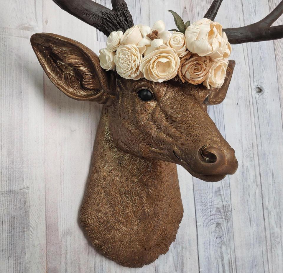 Faux taxidermy deer head that's been painted antique gold with chestnut brown accenting. The antlers are chestnut brown and atop the deers head is a cream floral crown made out of wooden sola flowers