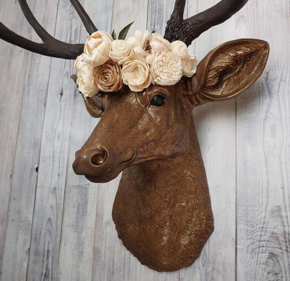 Faux taxidermy deer head that's been painted antique gold with chestnut brown accenting. The antlers are chestnut brown and atop the deers head is a cream floral crown made out of wooden sola flowers