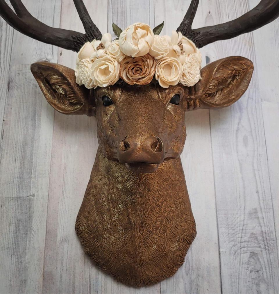 Faux taxidermy deer head that's been painted antique gold with chestnut brown accenting. The antlers are chestnut brown and atop the deers head is a cream floral crown made out of wooden sola flowers