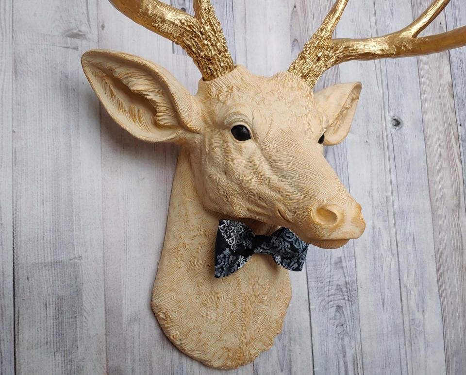 Faux taxidermy deer head painted cream with gold accenting and gold antlers. A fabric bow tie accents the neck, fabric color is dark blue multi with lighter blue and white
