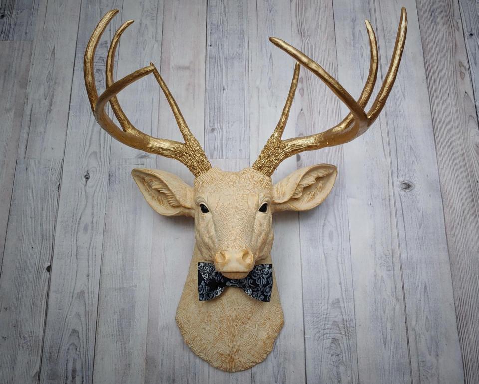 Faux taxidermy deer head painted cream with gold accenting and gold antlers. A fabric bow tie accents the neck, fabric color is dark blue multi with lighter blue and white.