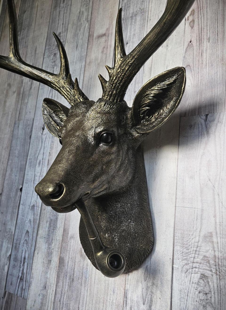 Rubbed bronze faux taxidermy deer head with gold accents. A smoking pipe extends from the deers mouth
