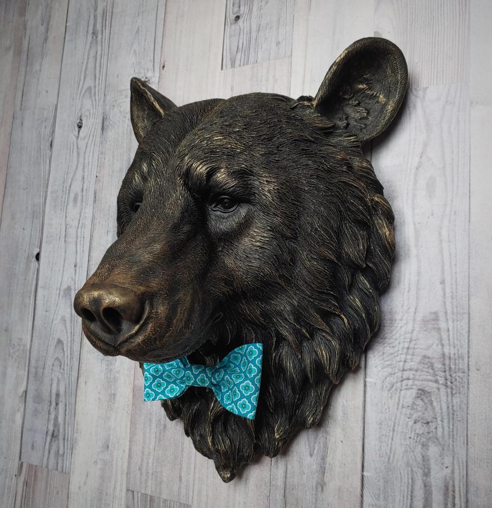 Faux taxidermy bear head wall mount, painted black with gold accenting. A turquoise and blue fabric bow tie accents the bears neck.
