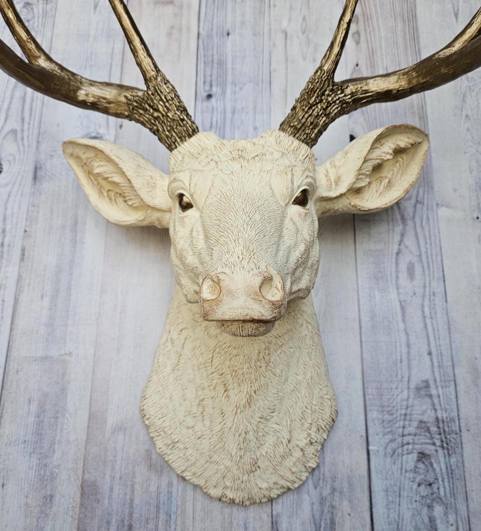Faux taxidermy deer head, painted antique white with antique gold accenting. The antlers are antique gold a small brass bird is perched on the left facing antler.