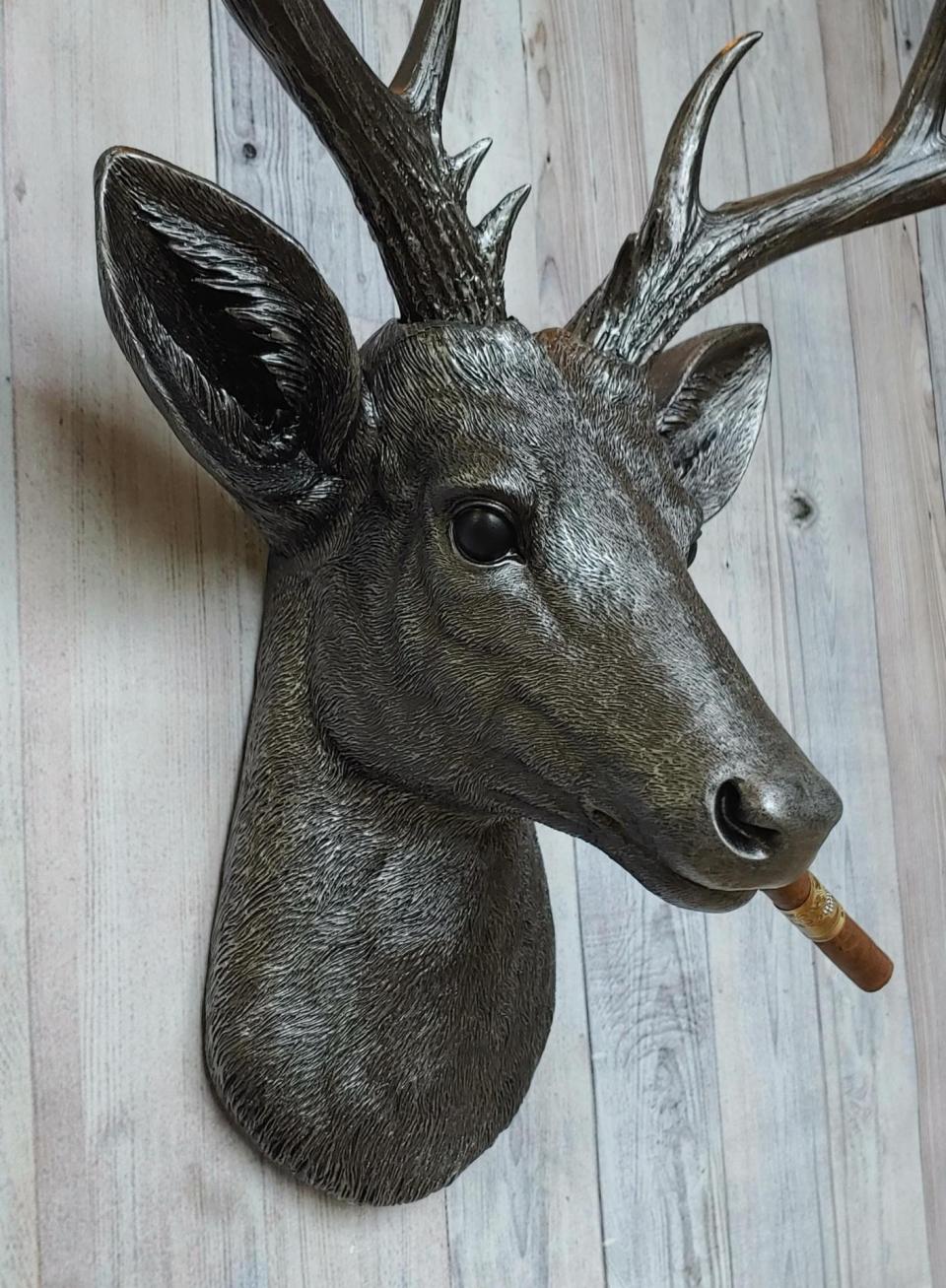 Faux taxidermy deer head painted rubbed bronze with rubbed bronze antlers and silver accents. A cigar is in the deers mouth.