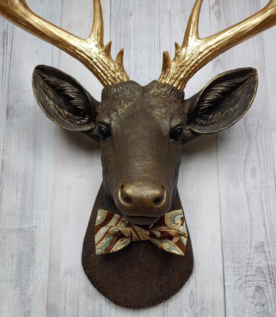 Faux taxidermy deer head, painted chestnut brown with gold accents. The antlers are gold and there is a multi colored brown tonal bow tie on the deers neck
