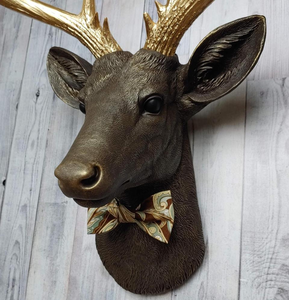 Faux taxidermy deer head, painted chestnut brown with gold accents. The antlers are gold and there is a multi colored brown tonal bow tie on the deers neck