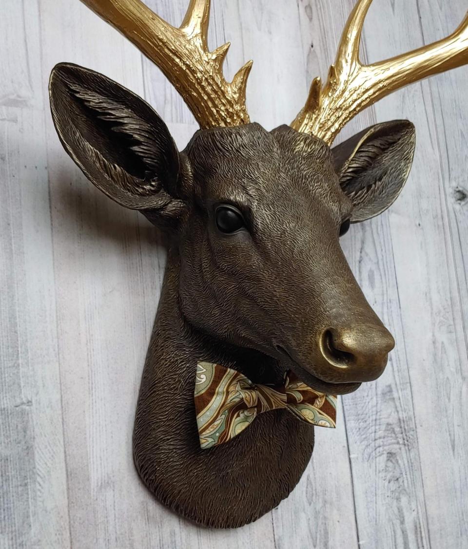 Faux taxidermy deer head, painted chestnut brown with gold accents. The antlers are gold and there is a multi colored brown tonal bow tie on the deers neck