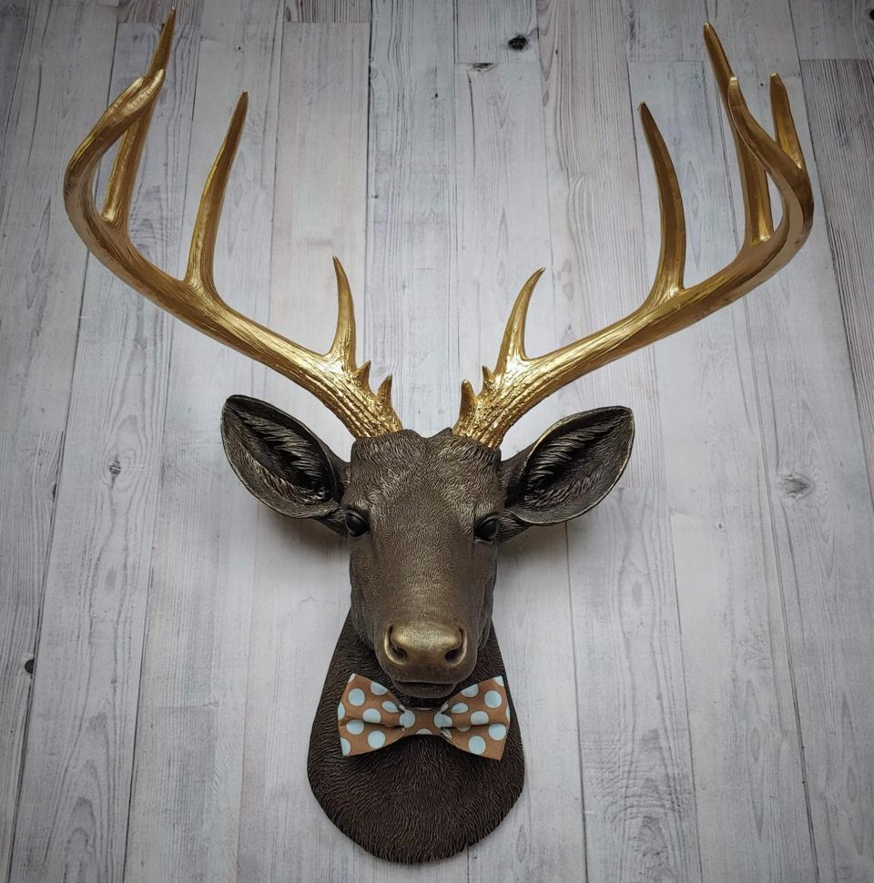 Faux taxidermy deer head. Painted chestnut brown with gold accents and gold antlers. There is a blue and brown polka dot bow tie on the deers neck.