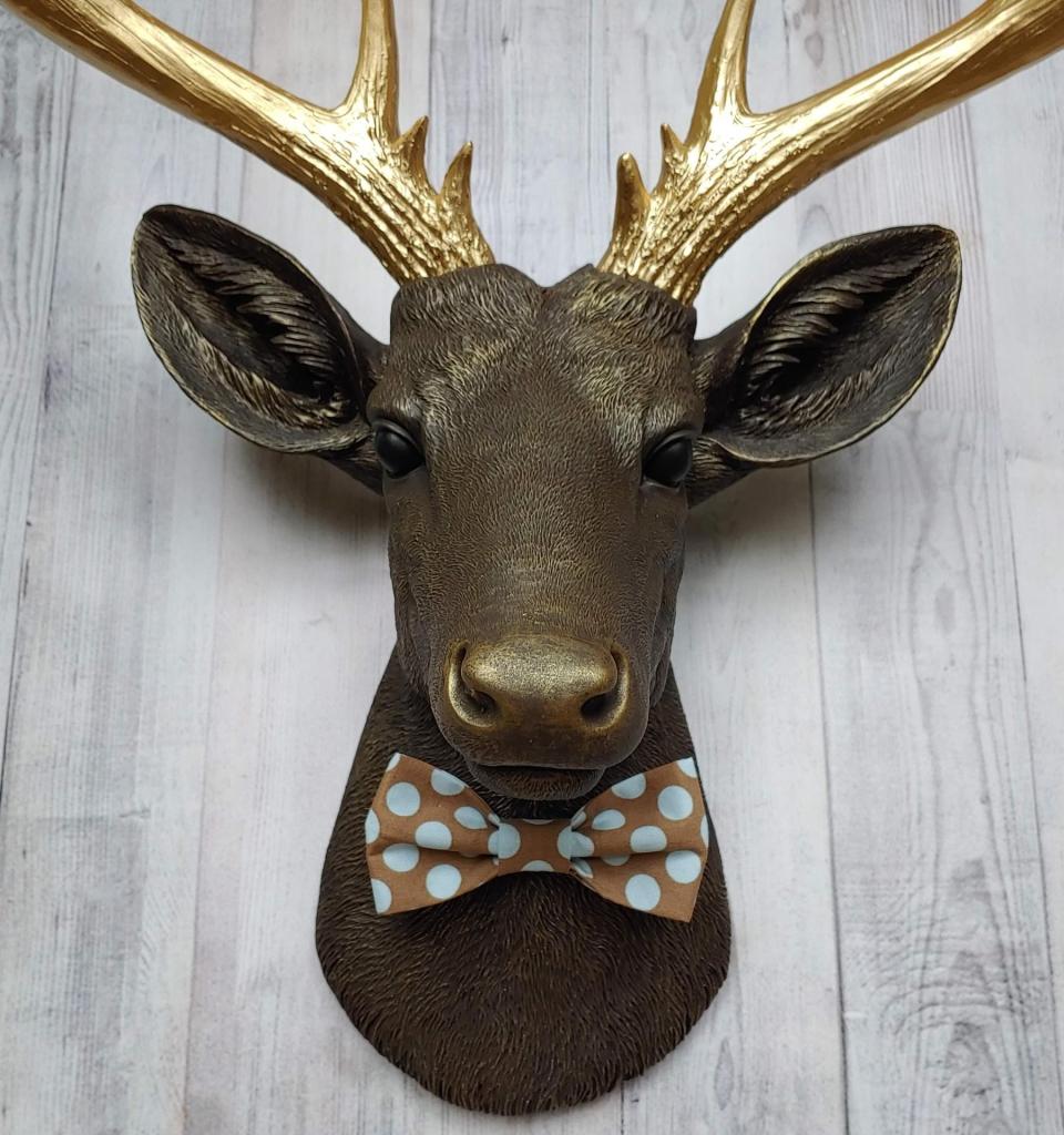 Faux taxidermy deer head. Painted chestnut brown with gold accents and gold antlers. There is a blue and brown polka dot bow tie on the deers neck.