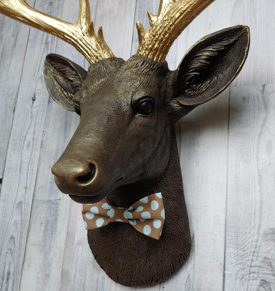 Faux taxidermy deer head. Painted chestnut brown with gold accents and gold antlers. There is a blue and brown polka dot bow tie on the deers neck.