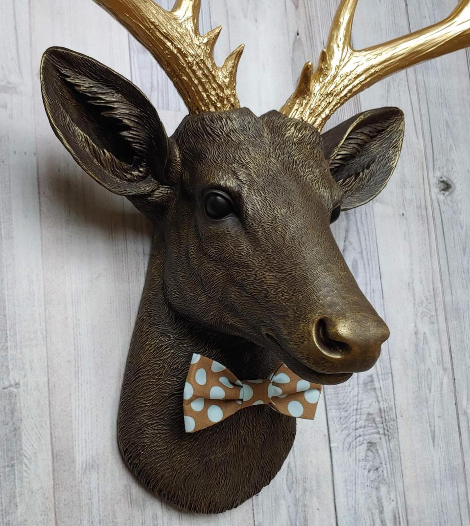 Faux taxidermy deer head. Painted chestnut brown with gold accents and gold antlers. There is a blue and brown polka dot bow tie on the deers neck.
