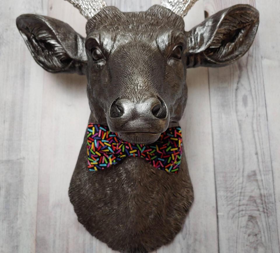 Faux taxidermy deer head painted rubbed bronze with silver accents and silver antlers. The deer has a bow tie on it's neck in black with colorful sprinkles.