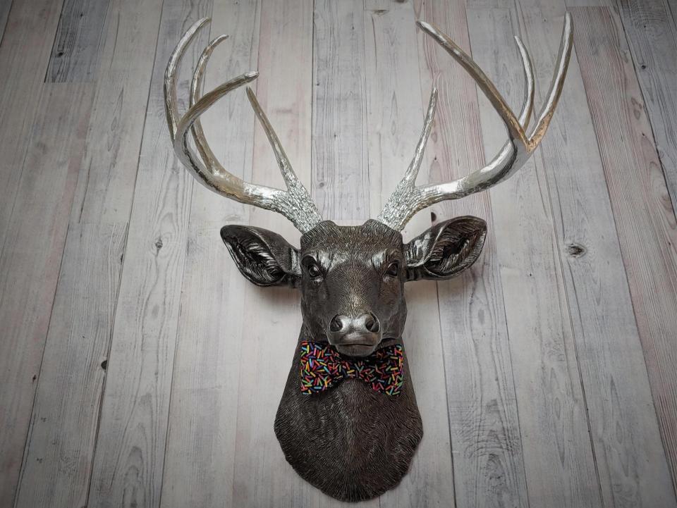Faux taxidermy deer head painted rubbed bronze with silver accents and silver antlers. The deer has a bow tie on it's neck in black with colorful sprinkles.