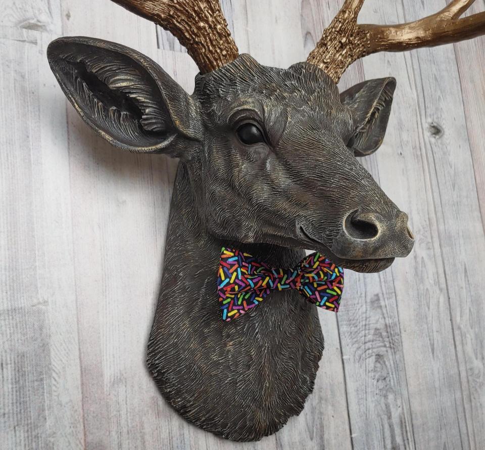 Faux taxidermy dee head painted chestnut brown and accented with antique white and antique gold. The antlers have been painted antique gold and the eyes have been painted black. A fabric bow tie that has a black background with multi colored sprinkles accents the deers neck.