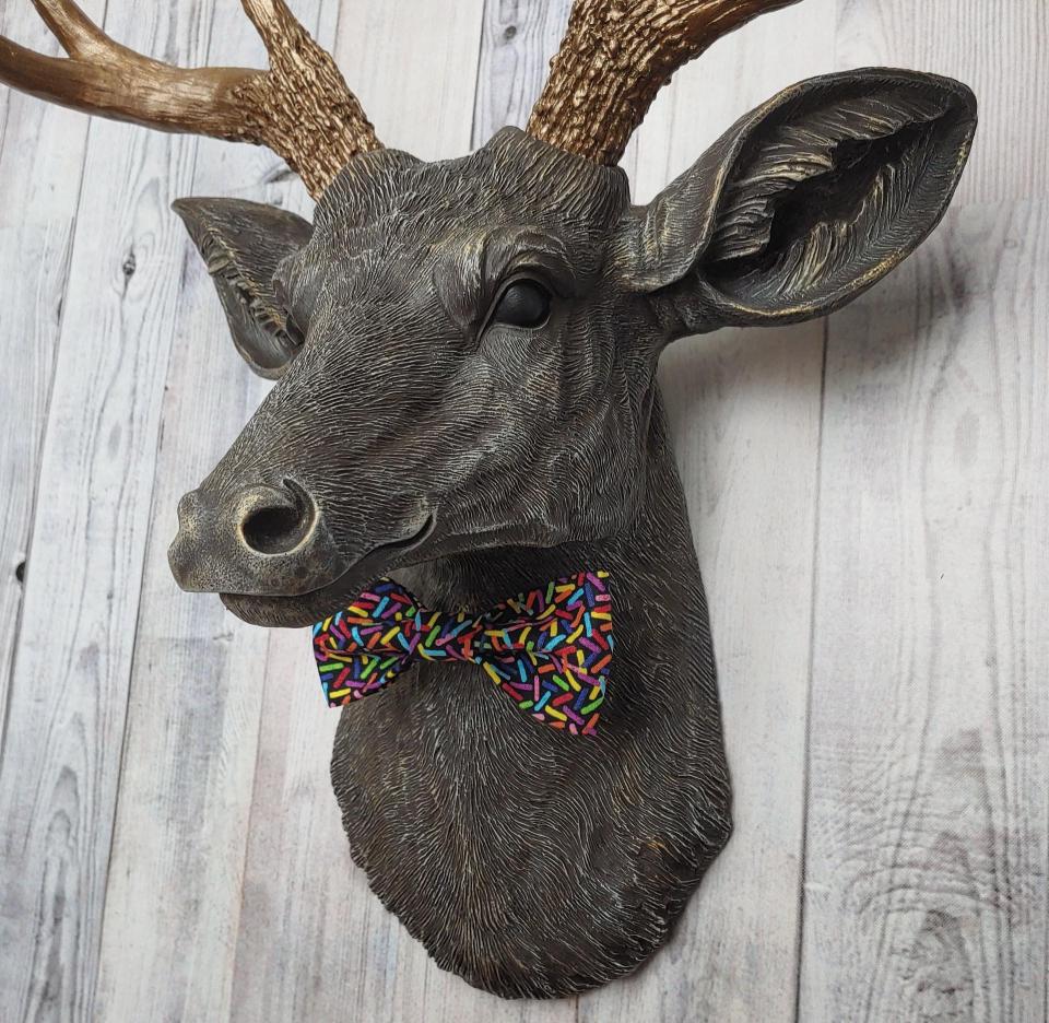 Faux taxidermy dee head painted chestnut brown and accented with antique white and antique gold. The antlers have been painted antique gold and the eyes have been painted black. A fabric bow tie that has a black background with multi colored sprinkles accents the deers neck.