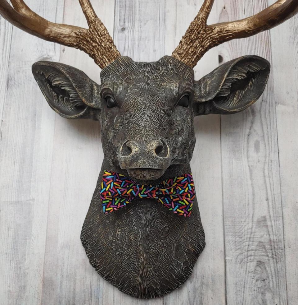 Faux taxidermy dee head painted chestnut brown and accented with antique white and antique gold. The antlers have been painted antique gold and the eyes have been painted black. A fabric bow tie that has a black background with multi colored sprinkles accents the deers neck.