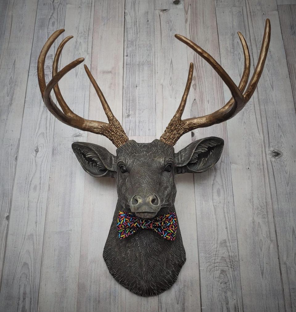 Faux taxidermy dee head painted chestnut brown and accented with antique white and antique gold. The antlers have been painted antique gold and the eyes have been painted black. A fabric bow tie that has a black background with multi colored sprinkles accents the deers neck.